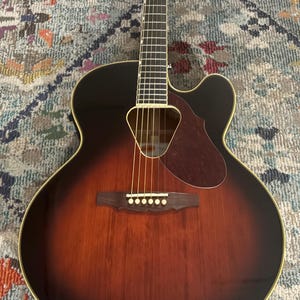 May include: Acoustic-electric guitar with a sunburst finish. The guitar features a dark brown body that fades to a lighter brown in the center, with a black headstock and a cutaway design. The soundhole is visible.