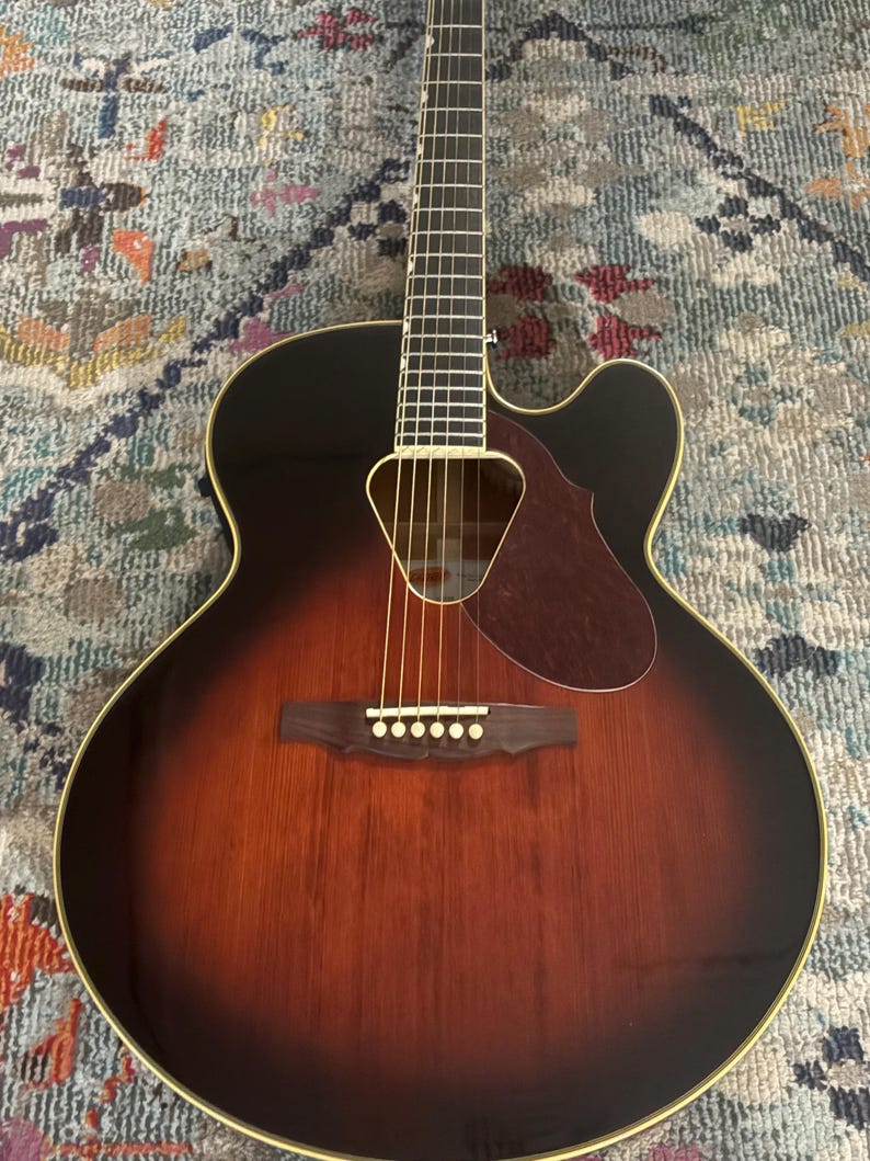 May include: Acoustic-electric guitar with a sunburst finish. The guitar features a dark brown body that fades to a lighter brown in the center, with a black headstock and a cutaway design. The soundhole is visible.