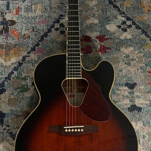May include: Acoustic-electric guitar with a sunburst finish, featuring a black top and a brown body. The guitar has a cutaway design, a pickguard, and six strings. The headstock has the brand name.