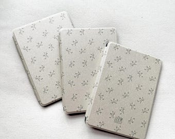 May include: Three white book covers with a small floral pattern and a small logo on the bottom right corner.