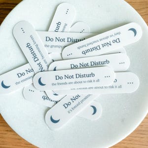May include: A collection of white, oval-shaped tags with the words "Do Not Disturb" printed in a dark gray font. Each tag features a crescent moon graphic and a short phrase. The tags are arranged on a white, marble-like plate.