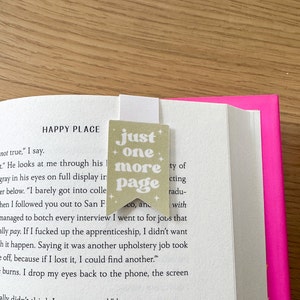 May include: A beige bookmark with the text "just one more page" in a whimsical font. The bookmark has a star design on each corner and is placed on a book with a pink cover.