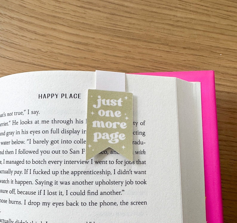 May include: A beige bookmark with the text "just one more page" in a whimsical font. The bookmark has a star design on each corner and is placed on a book with a pink cover.