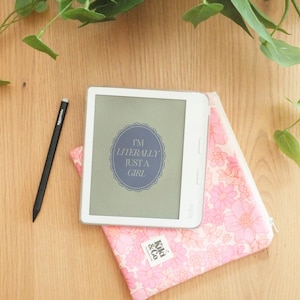 Könnte beinhalten: Ein weißer Kobo E-Reader mit dem Text „I'M LITERALLY JUST A GIRL“ in einem blauen Rahmen. Er liegt auf einem rosa Reißverschlussetui mit Blumenmuster. Ein schwarzer Stift befindet sich links.