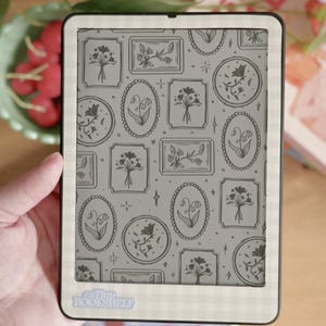 May include: An e-reader with a floral pattern on the screen, held in a hand. The screen displays various framed floral illustrations in gray tones. The e-reader has a white border with a gingham pattern. The text "Tiny Bookshelf" is visible on the bottom.