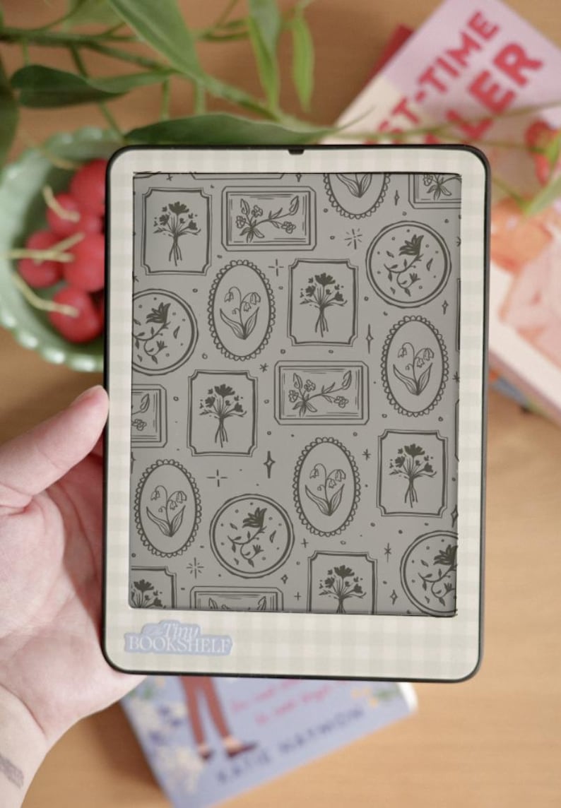 May include: An e-reader with a floral pattern on the screen, held in a hand. The screen displays various framed floral illustrations in gray tones. The e-reader has a white border with a gingham pattern. The text "Tiny Bookshelf" is visible on the bottom.