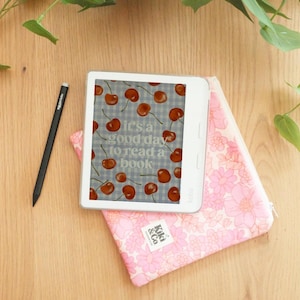 Pode incluir: Um e-reader branco com um padrão de cerejas e o texto "it's a good day to read a book" está em cima de uma capa floral rosa. Uma caneta stylus preta está em uma superfície de madeira ao lado do e-reader e da capa.