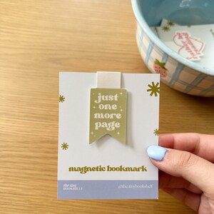 May include: A gold magnetic bookmark with the text "just one more page" in a retro font. The bookmark is on a white card with gold stars and the text "magnetic bookmark", "the tiny bookshelf", and "@the.tinybookshelf".