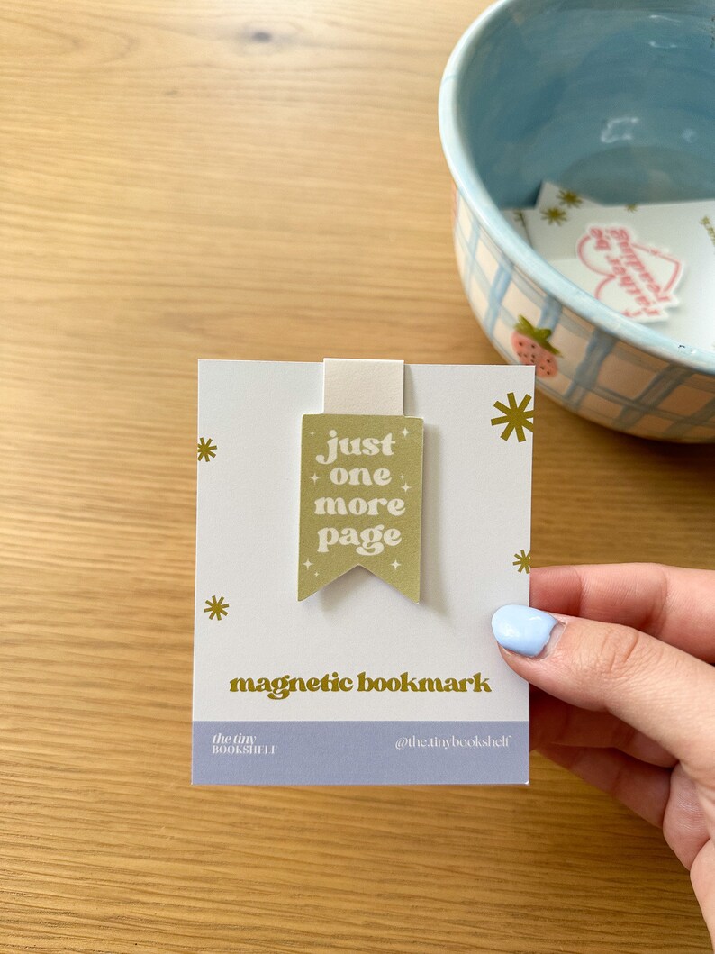 May include: A gold magnetic bookmark with the text "just one more page" in a retro font. The bookmark is on a white card with gold stars and the text "magnetic bookmark", "the tiny bookshelf", and "@the.tinybookshelf".