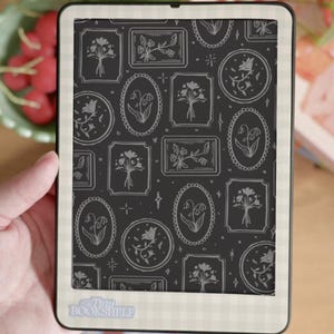 May include: An e-reader displays a floral pattern on its screen. The pattern features various framed floral designs in white against a dark background. The e-reader is held in a hand, with a book and a bowl of red berries in the background. The text "Tiny Bookshelf" is visible.
