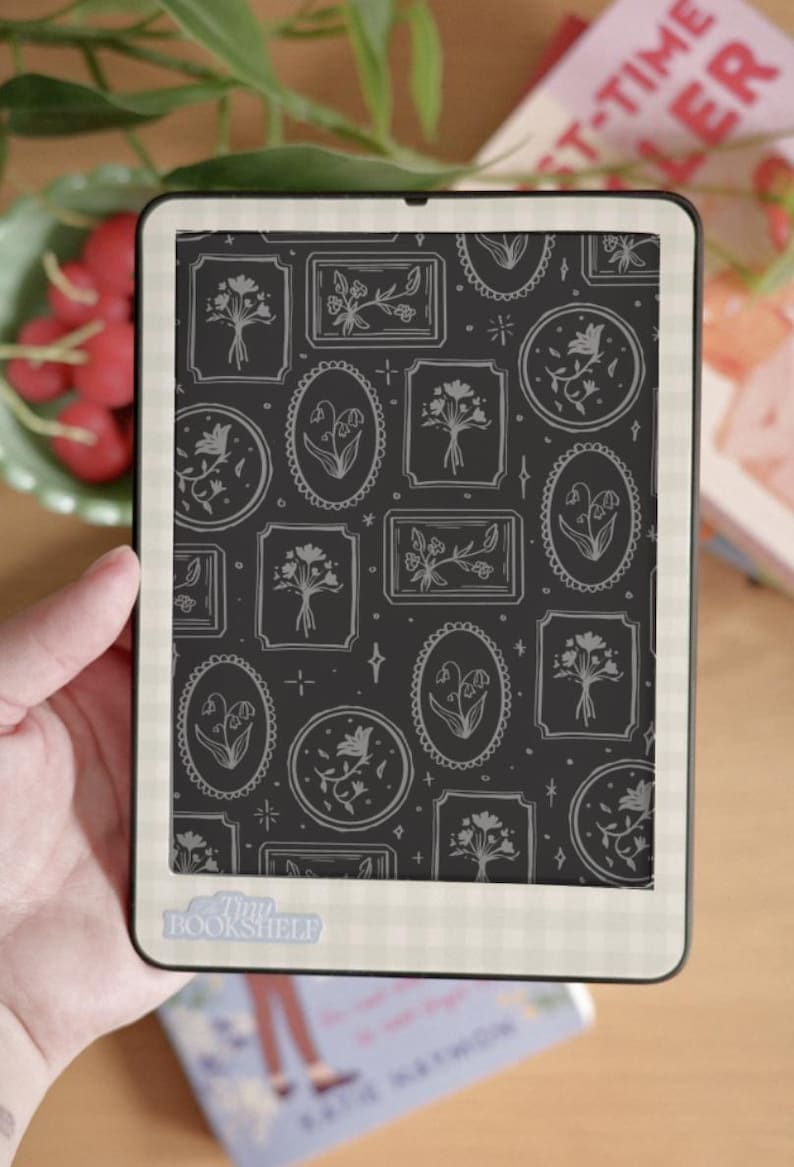 May include: An e-reader displays a floral pattern on its screen. The pattern features various framed floral designs in white against a dark background. The e-reader is held in a hand, with a book and a bowl of red berries in the background. The text "Tiny Bookshelf" is visible.