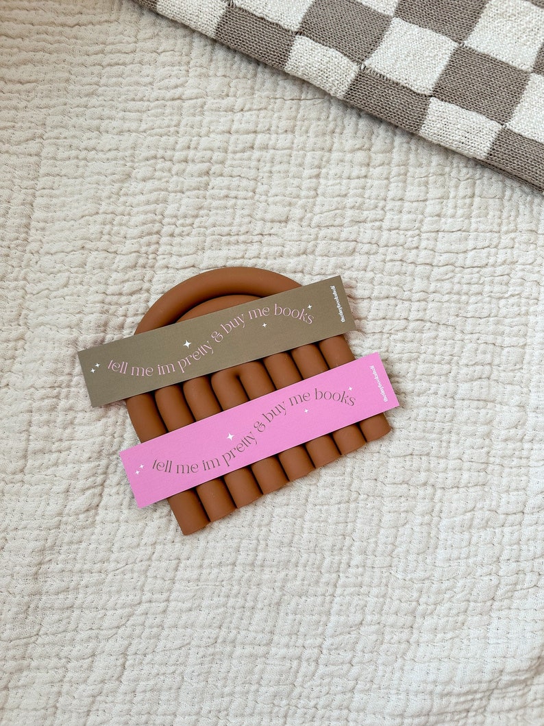 May include: Two brown silicone bookmarks with the text "tell me im pretty & buy me books" in pink and gray lettering.