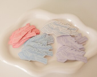 May include: A close-up of a pile of pastel-colored stickers with the text "Midnight Readers Society" on them. The stickers are in the shape of a book with a ribbon bookmark.