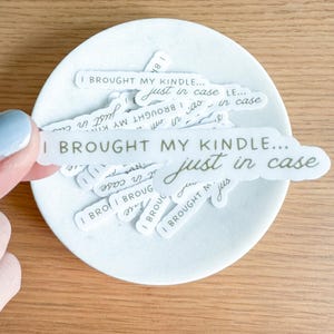 May include: A hand holds a white sticker with the text "I BROUGHT MY KINDLE... just in case" over a white, round dish. Other similar stickers are in the dish. The stickers have a handwritten font in a light gray color.