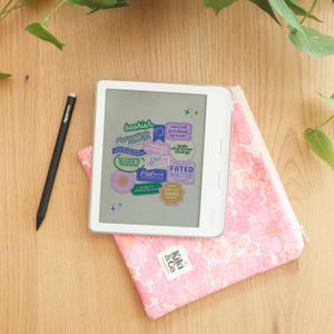 Könnte beinhalten: Ein weißer Kobo-E-Reader mit Aufklebern mit der Aufschrift „bookish“ und „Fated Mates“ liegt auf einer rosa Blumentasche. Ein schwarzer Stift und grüne Blätter sind ebenfalls sichtbar. Die Hülle hat einen Reißverschluss und den Text „Kiki & Co.“
