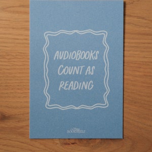 May include: A blue card with white text that reads "Audiobooks count as reading" in a decorative frame. The bottom of the card has the text "The Bookshelf" in white.