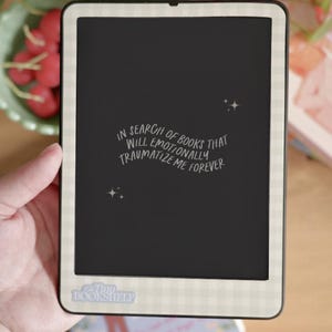 Puede incluir: Un lector electrónico negro con un borde a cuadros blanco y gris. La pantalla muestra el texto "In search of books that will emotionally traumatize me forever." El lector electrónico tiene una pegatina que dice "Tiny Bookshelf".