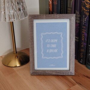 May include: A framed print with a light blue background and white text that reads "It's okay to take a break". The print is in a wooden frame with a gray finish. The print is sitting on a wooden surface.