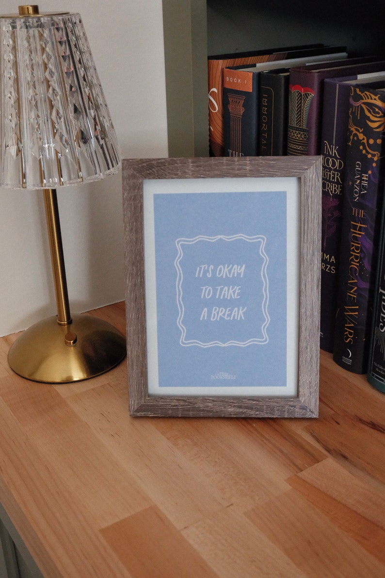May include: A framed print with a light blue background and white text that reads "It's okay to take a break". The print is in a wooden frame with a gray finish. The print is sitting on a wooden surface.