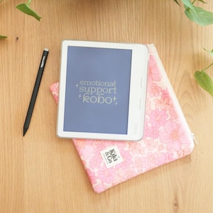 Op de afbeelding: Een witte Kobo e-reader met de tekst "emotional support kobo" op het scherm, rustend op een roze bloemenpatroon hoes. Een zwarte stylus en een roze bloemenhoes met het "Kiki & Co" logo zijn ook aanwezig op een houten oppervlak.