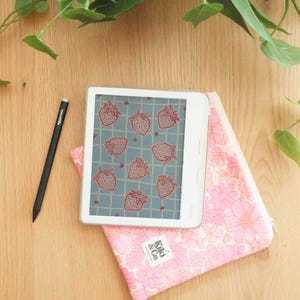 May include: A white e-reader displaying a strawberry pattern sits on a pink floral pouch. A black stylus and a pink floral pouch with the text "Kiki & Co" are also present. The items are on a wooden surface.