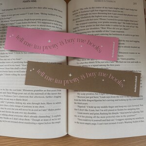 May include: Two pink bookmarks with the text "tell me im pretty & buy me books" printed on them. The bookmarks are placed on an open book with text visible on the pages.