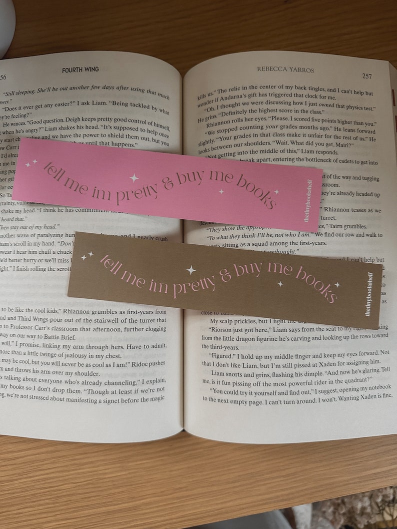 May include: Two pink bookmarks with the text "tell me im pretty & buy me books" printed on them. The bookmarks are placed on an open book with text visible on the pages.