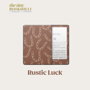 May include: A brown and white Kindle e-reader with a brown horseshoe pattern cover. The text "Rustic Luck" is printed below the e-reader. The text on the e-reader screen reads "Chapter 1" and includes a short excerpt from a book.