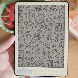 Op de afbeelding: Een zwart-witte e-reader met een grijs scherm waarop een patroon van verschillende illustraties te zien is, waaronder een eend, een konijn, een boek, een kopje koffie, een strik, een kers en een croissant. De e-reader heeft een witte rand met een geruit patroon en de tekst "tiny bookshelf" onderaan.