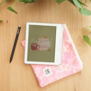 Puede incluir: Un lector electrónico Kobo blanco con la inscripción "Cozy Readers Club" en la pantalla, un lápiz óptico negro y una funda floral rosa. La funda tiene una etiqueta "Kiki & Co". Los artículos están sobre una superficie de madera con vegetación.