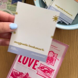 May include: A pink and white book with the title "LOVE" and the text "ROSE HILL SERIES" on the cover. The book is a USA Today Bestselling Author book by Elsie Silver. The book is titled "revelation!" by Ali Hazelwood. A hand is holding a light blue and white magnetic bookmark with the text "magnetic bookmark" on it.