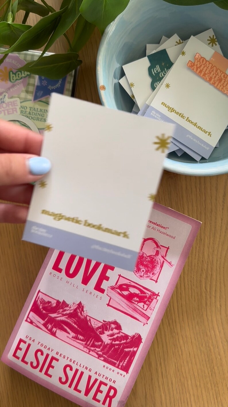 May include: A pink and white book with the title "LOVE" and the text "ROSE HILL SERIES" on the cover. The book is a USA Today Bestselling Author book by Elsie Silver. The book is titled "revelation!" by Ali Hazelwood. A hand is holding a light blue and white magnetic bookmark with the text "magnetic bookmark" on it.
