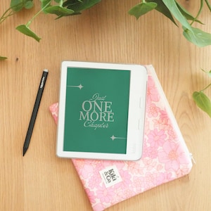Op de afbeelding: Een witte e-reader met een groen scherm met de tekst "Just One More Chapter", rustend op een roze bloemenetui. Een zwarte stylus en groene bladeren zijn ook zichtbaar op een houten oppervlak.