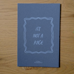 May include: A dark gray card with white text that reads "It's not a race" inside a decorative frame. The text "The Bookshelf" is printed in white at the bottom of the card.