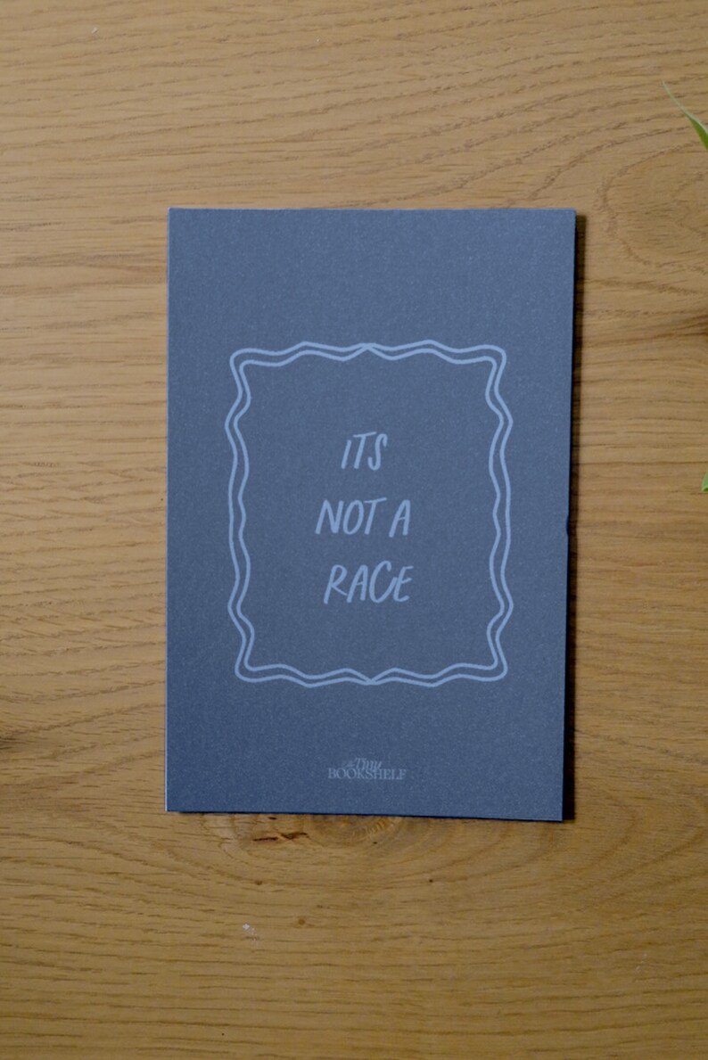 May include: A dark gray card with white text that reads "It's not a race" inside a decorative frame. The text "The Bookshelf" is printed in white at the bottom of the card.