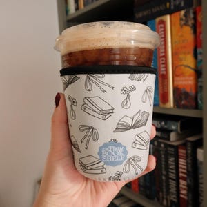 May include: A white coffee cup sleeve with a black trim and a pattern of books, flowers, and ribbons. The text "Tiny Book Shelf" is printed in blue on the sleeve.