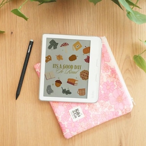 May include: A white e-reader with a screen displaying autumn-themed illustrations and the text "IT'S A GOOD DAY To Read." It rests on a pink floral case with a matching stylus. The case has a zipper and a label that says "Kiki & Co."