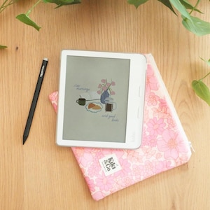 以下が含まれることがあります： 花柄のイラストと「slow mornings and good books」の文字が表示された白いKobo電子書籍リーダー。ピンクの花柄ケースの上に置かれ、「Kiki & Co」のラベルが付いています。黒いスタイラスペンも写っています。