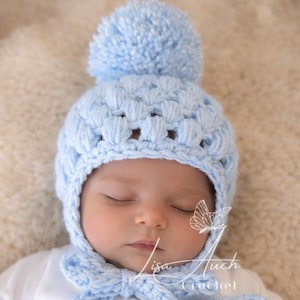 May include: A light blue crochet baby bonnet with a matching pom-pom and tie closure. The bonnet is made of soft yarn and features a textured stitch pattern. The text "Lisa Auch Crochet" is visible.