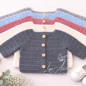 Op de afbeelding: Een stapel handgehaakte babyvesten in verschillende kleuren: grijs, lichtblauw, roze en crème. Elk vest heeft houten knopen en lange mouwen. De afbeelding bevat babyschoenen, een knuffel en een houten bijtring.