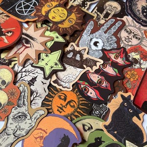 May include: A selection of stickers with an occult theme. The stickers feature illustrations of suns, moons, eyes, bats, cats, hands, and stars in various colours including orange, brown, green, and black. The stickers have a vintage, handmade aesthetic.