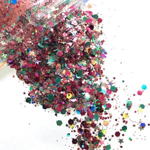 May include: A close-up of a pile of glitter in various shapes and colours. The glitter includes stars, hexagons, and other shapes in shades of pink, green, and copper. The glitter is scattered on a white surface.