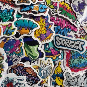 May include: A collection of assorted vinyl stickers featuring vibrant graffiti-style artwork, cartoon characters, and stylised lettering. The designs include bright colours like neon pink, blue, yellow, and green, with text elements such as STREET and SIMPLY FUN.