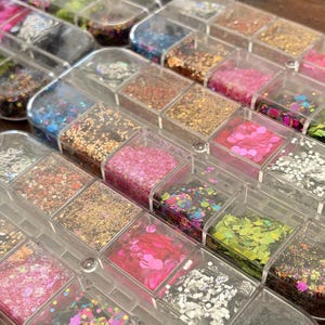 May include: A clear plastic organiser filled with various colours and shapes of glitter. The glitter includes gold, silver, pink, green, and black, in a variety of sizes and textures. The compartments are arranged in rows and columns.