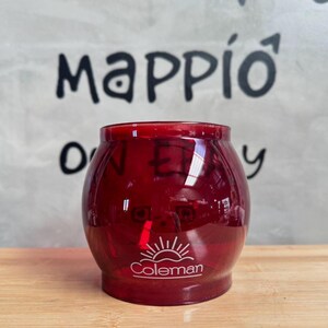 May include: A transparent, deep red Coleman camping lantern with a rounded shape. The Coleman logo is visible at the bottom. The background features the text "P&P Camping" and "mappio".