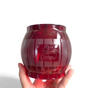 May include: A red Pyrex Coleman glass jar with a barrel design. The jar is made in the USA and has the Coleman logo. The jar is held by a hand against a white background.