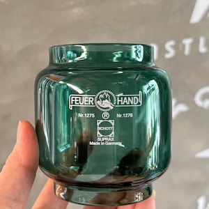 Puede incluir: Un tarro de vidrio verde oscuro con un cuerpo redondeado y un borde ligeramente ensanchado. El tarro tiene el texto "FEUER HAND" y "SCHOTT SUPRAX Made in Germany" impreso en la parte delantera. El tarro está en una mano.