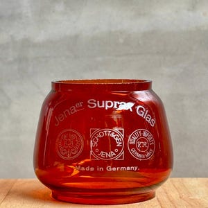 Puede incluir: Un tarro de vidrio rojo con el texto "Jenaer Suprax Glas" y "Made in Germany." El tarro tiene tres logotipos circulares, uno con el texto "Schott & Gen Jena" y otro con el texto "Quality-Imports F.R. German Make."