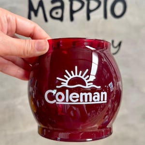 May include: A round, deep red glass mate cup featuring a white graphic of a rising sun above the word Coleman printed on the front. The vessel has a smooth, glossy finish with a wide opening and a slightly narrower base, designed for drinking.