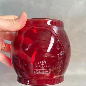 May include: A vintage, ruby red Pyrex glass jar for Coleman, Made in U.S.A. The jar has a rounded shape with a slightly flared base. The Pyrex and Coleman logos are visible on the front.